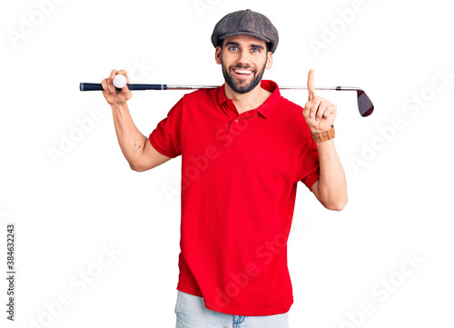 Young handsome man with beard playing golf holding club and ball surprised with an idea or question pointing finger with happy face, number one