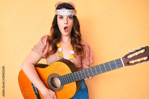 Young beautiful brunette woman wearing boho style playing classical guitar scared and amazed with open mouth for surprise, disbelief face photo