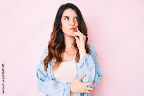 Young beautiful brunette woman wearing casual clothes over pink background serious face thinking about question with hand on chin, thoughtful about confusing idea