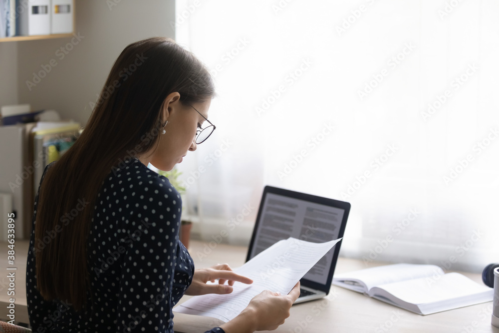 Back rear view focused young woman in eyeglasses reading paper document, checking electronic report on computer. Concentrated smart ambitious businesswoman preparing research, freelance work. Back rear view focused young woman in eyeglasses reading paper document, checking electronic report on computer. Concentrated smart ambitious businesswoman preparing research, freelance work.