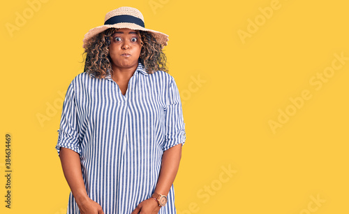 Young african american plus size woman wearing summer hat puffing cheeks with funny face. mouth inflated with air, crazy expression.