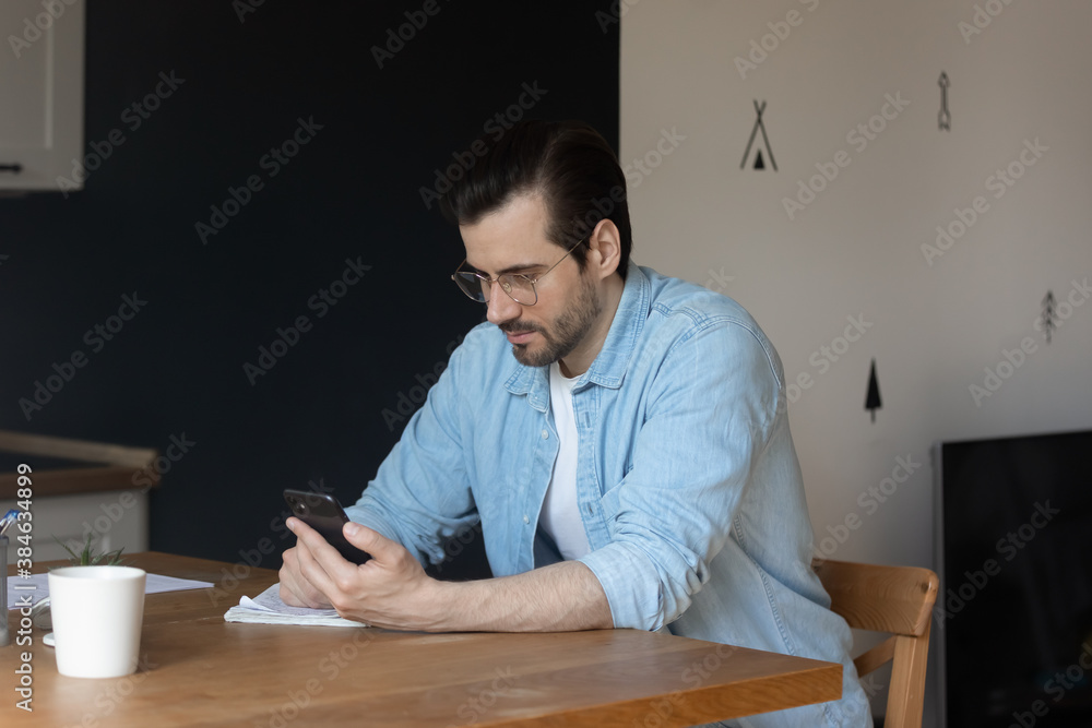 Focused young man using smartphone, sitting at home office table. Serious 30s guy in eyeglasses web surfing information or involved in communication with colleagues, distracted from work indoors. Focused young man using smartphone, sitting at home office table. Serious 30s guy in eyeglasses web surfing information or involved in communication with colleagues, distracted from work indoors.