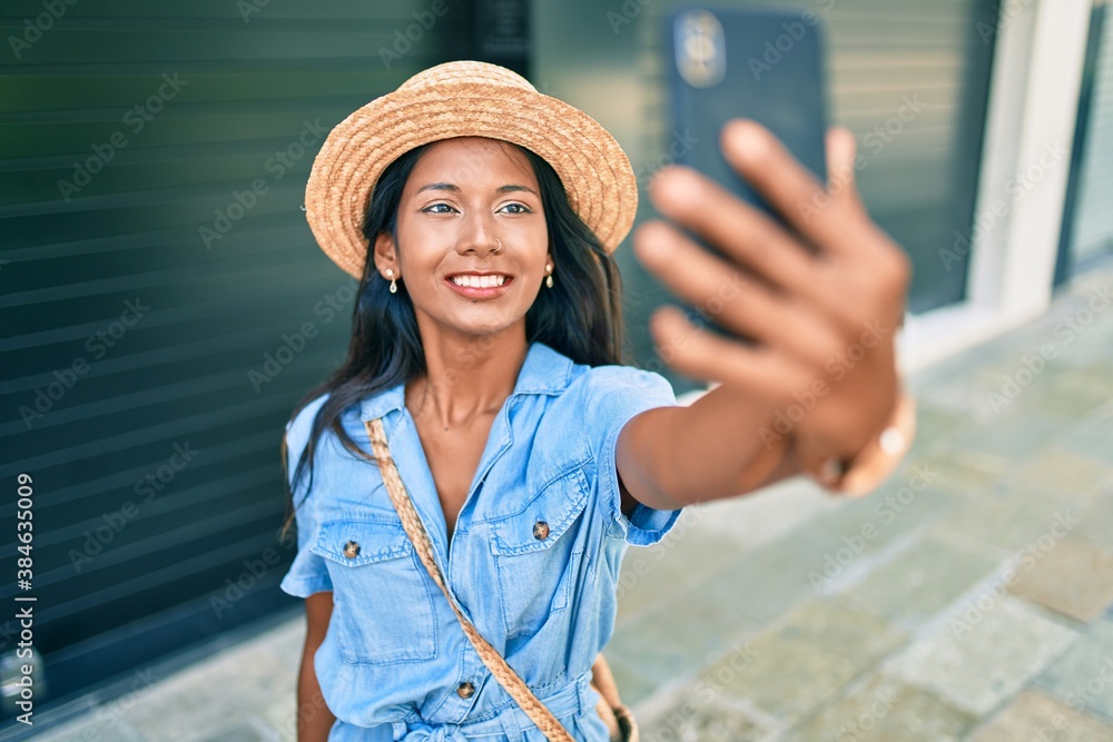 Young beautiful indian woman wearing summer hat smiling happy making selfie by the smartphone at the city.