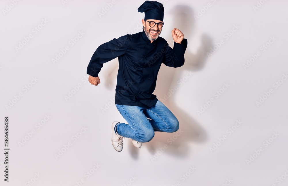 Middle age handsome hispanic man wearing glasses and cook uniform smiling happy. Jumping with smile on face celebrating with fists up over isolated white background