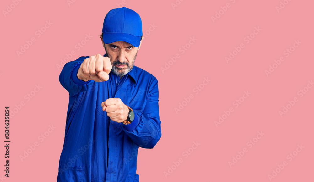 Middle age handsome man wearing mechanic uniform punching fist to fight, aggressive and angry attack, threat and violence