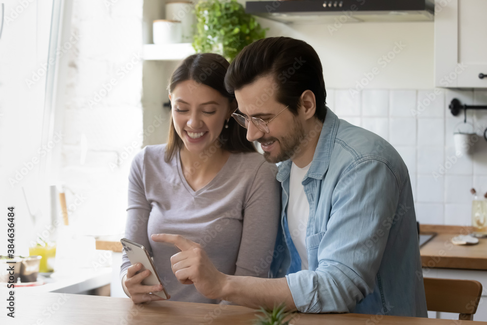 Obraz premium Pretty young woman holding smartphone in hands, showing funny photo editing application or online game to smiling handsome man, using electronic device together at free leisure time at home.