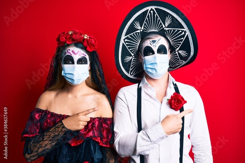 Couple wearing day of the dead costume wearing medical mask smiling and looking at the camera pointing with two hands and fingers to the side.
