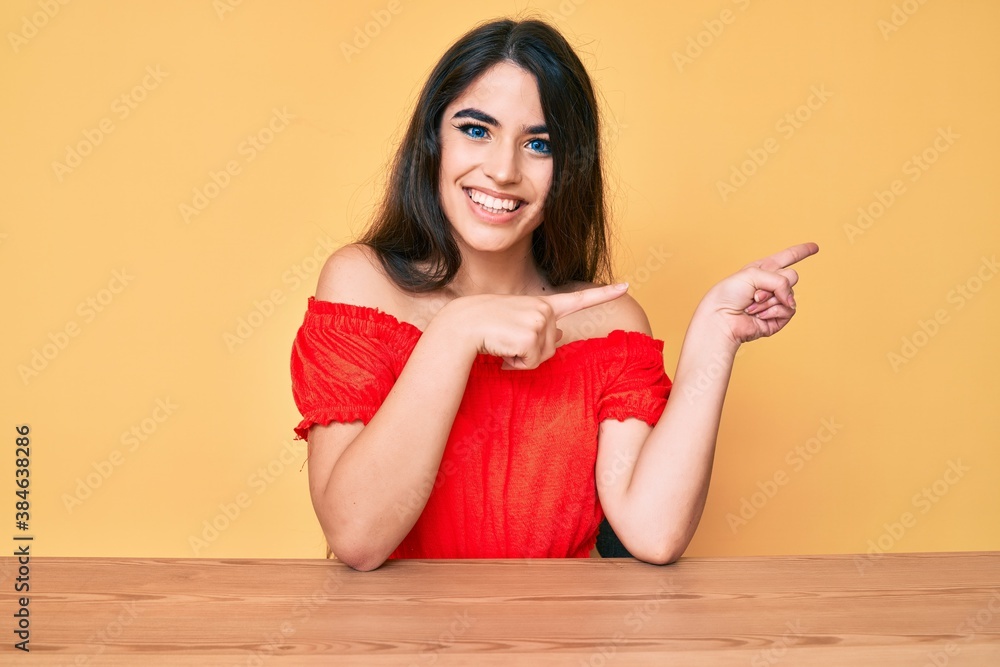 Brunette teenager girl wearing casual clothes sitting on the table smiling and looking at the camera pointing with two hands and fingers to the side. Brunette teenager girl wearing casual clothes sitting on the table smiling and looking at the camera pointing with two hands and fingers to the side.