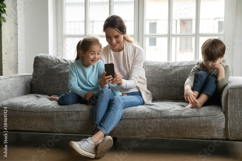 Smiling mom and little daughter have fun using modern cellphone gadget, offended hurt son child sit separately feel lonely abandoned. Happy mother spend weekend with one kid. Family problem concept.