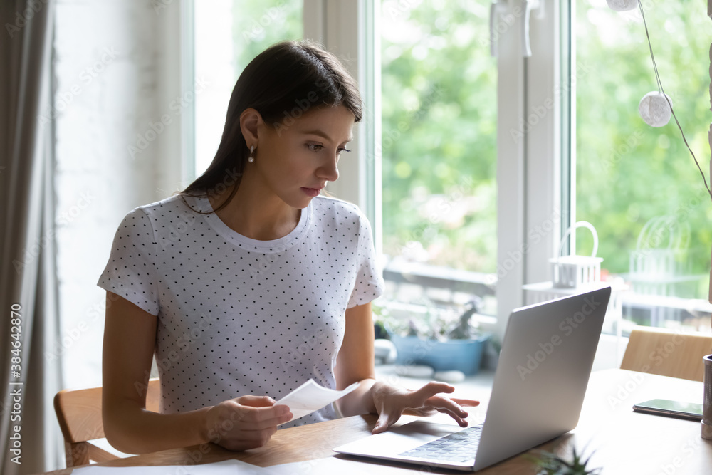 Thoughtful concentrated young woman holding paper bills in hands, entering payment information in computer application, paying utility expenditures online using laptop software alone at home. Thoughtful concentrated young woman holding paper bills in hands, entering payment information in computer application, paying utility expenditures online using laptop software alone at home.