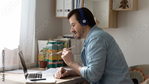 Focused young businessman in glasses wearing modern wireless headphones, involved in video conference call communication discussing project ideas with colleagues or partners remotely at home office.