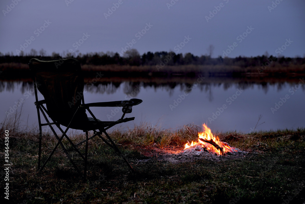 Tourist chair, folding chair, bonfire on the river bank in late evening, twilight in the fall at sunset, relaxation and rest, early autumn, close-up Tourist chair, folding chair, bonfire on the river bank in late evening, twilight in the fall at sunset, relaxation and rest, early autumn, close-up