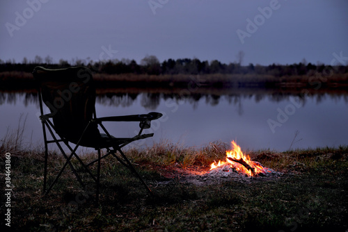Fototapeta Naklejka Na Ścianę i Meble -  Tourist chair, folding chair, bonfire on the river bank in late evening, twilight in the fall at sunset, relaxation and rest, early autumn, close-up