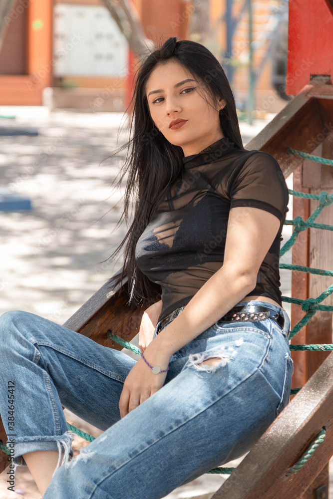 beautiful young latin woman with long straight hair sitting on rope, wearing a transparent blouse with jeans, outdoor photo with sun and wind beautiful young latin woman with long straight hair sitting on rope, wearing a transparent blouse with jeans, outdoor photo with sun and wind