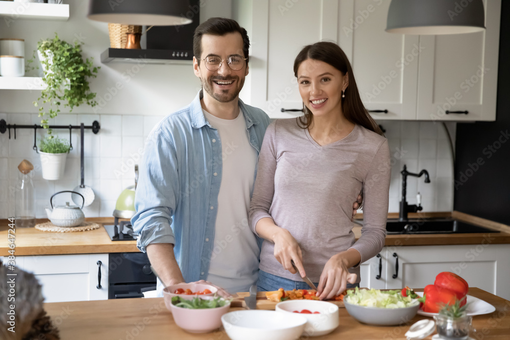 Portrait of happy young married couple chopping fresh vegetables at wooden board together in kitchen. Smiling beautiful woman enjoying cooking healthy food at free weekend time with handsome husband.