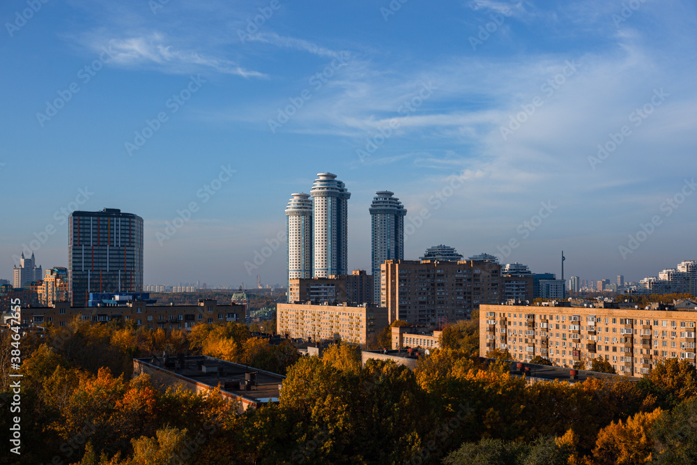 Fototapeta premium Urban landscape of new Moscow. Urban area with old and new housing during sunny day with foggy clouds and blue sky. In foreground are autumncolored treetops. Contrasty scene in architecture and light.