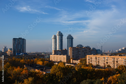 Urban landscape of new Moscow. Urban area with old and new housing during sunny day with foggy clouds and blue sky. In foreground are autumncolored treetops. Contrasty scene in architecture and light.