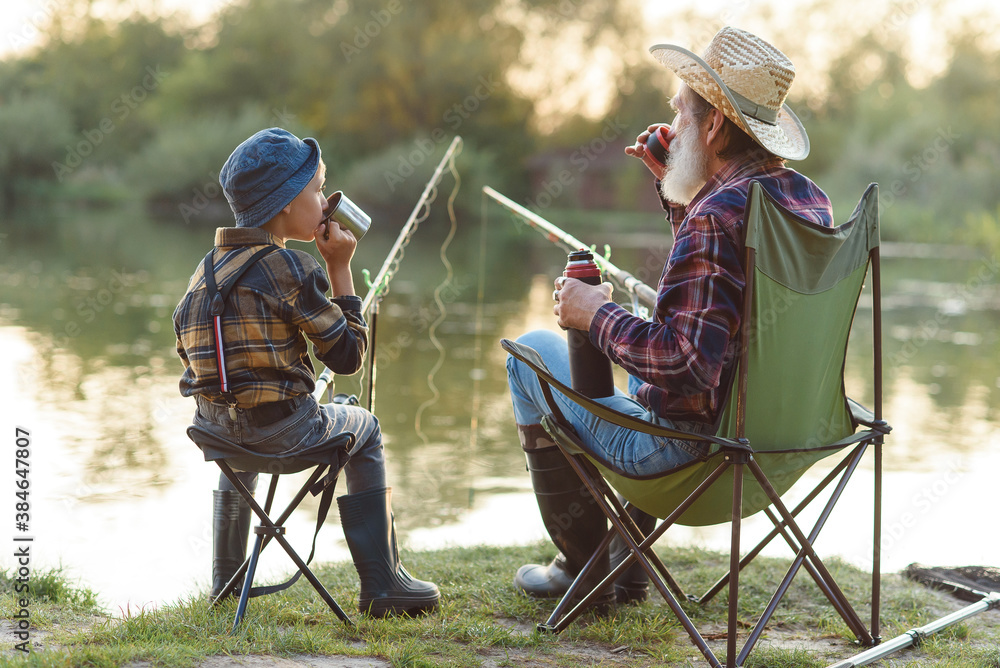 Careful grandpa with grey beard in straw hat sitting on folding chairs with  his grandchild on fishing and drinking tea. Stock Photo | Adobe Stock