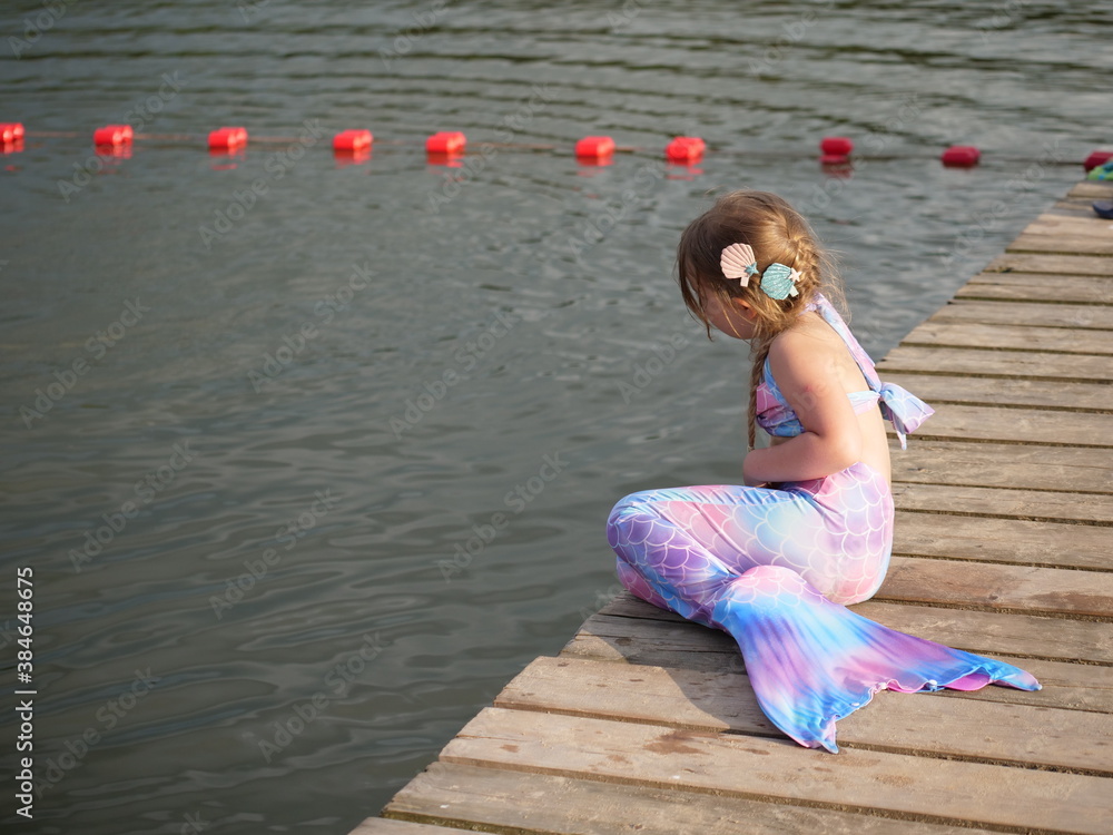Dziewczynka przebrana za syrenkę siedząca na pomoście nad brzegiem jeziora - obrazy, fototapety, plakaty 