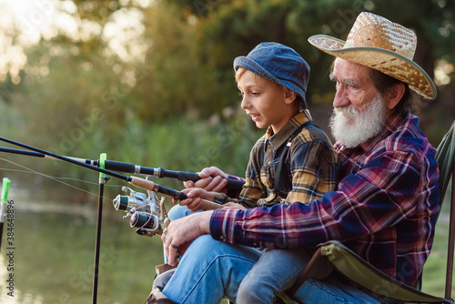Likable respected 70-aged bearded grandfather holding on knees 10-aged cute grandson and teaching him to catch fish.