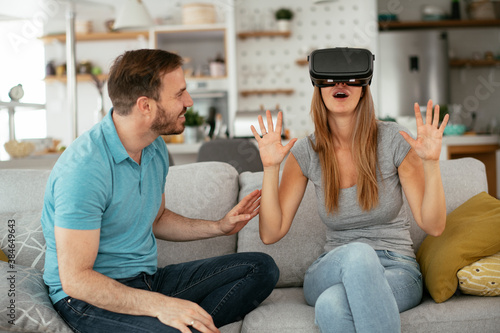 Smiling young woman using VR headset at home on couch. Woman and her husband enjoying virtual reality in apartment