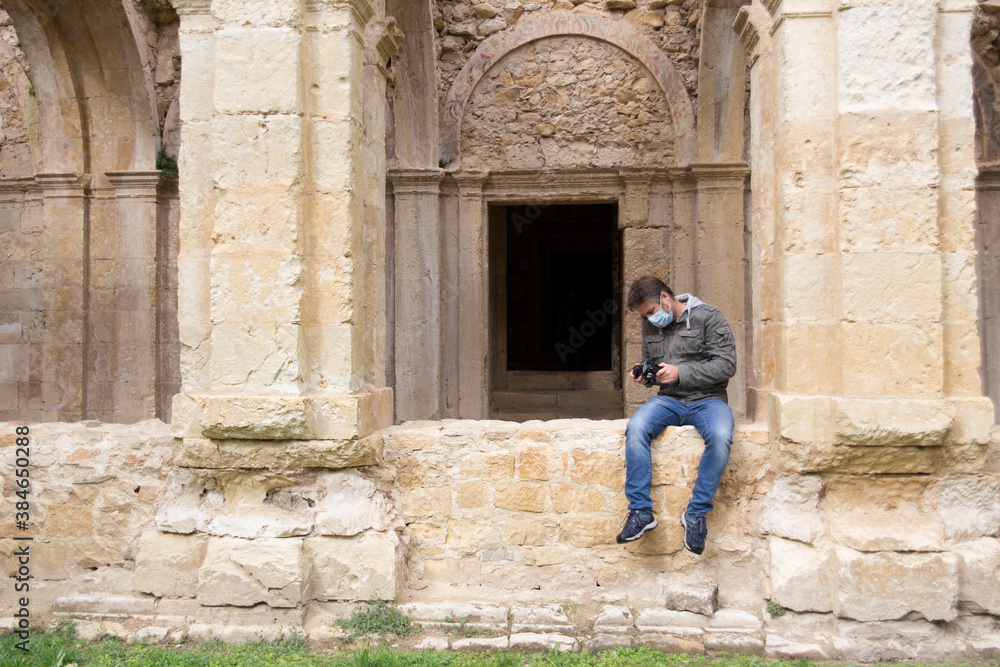 Joven sentado en un muro de una iglesia en ruinas con mascarilla higiénica y mirando su cámara de fotos