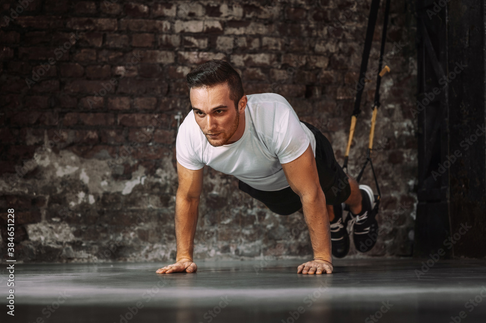 Muscular sportsman doing plank exercise with TRX ropes during suspension workout against shabby wall in gym Muscular sportsman doing plank exercise with TRX ropes during suspension workout against shabby wall in gym