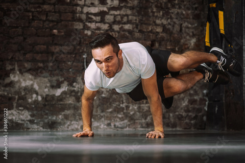 Strong male athlete with legs in suspension ropes breathing and doing exercise against grungy wall in gym