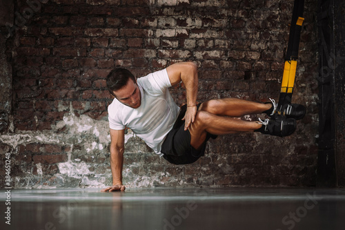 Powerful sportsman balancing on arm while doing exercise with TRX ropes against rough brick wall in gym