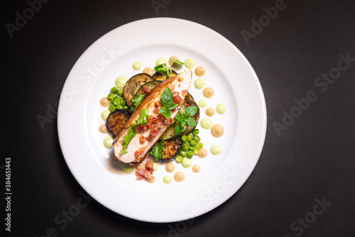 Turkey breast meat with fried pieces of bacon sits on baked eggplant and green peas aside, young and small basil leaves and cauliflower puree and pea puree. On a white plate and a black background.