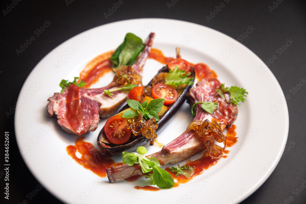 Lamb ribs with baked eggplant, cherry tomato wedges, caramelized onions, small basil leaves, micro greens, with red sauce on a white plate on a dark black background. Side view. Lamb ribs with baked eggplant, cherry tomato wedges, caramelized onions, small basil leaves, micro greens, with red sauce on a white plate on a dark black background. Side view.