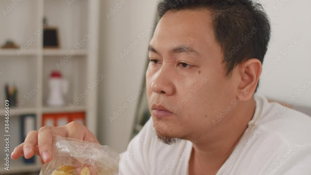 Asian man enjoys eating casava or potato chips while sitting on sofa at living room Asian man enjoys eating casava or potato chips while sitting on sofa at living room