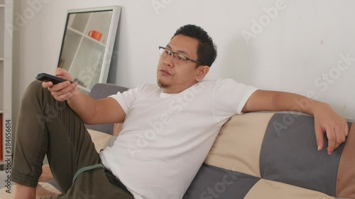 Bored tired  young Asian man sitting on the sofa and watching TV. Trying to choose interesting TV channel