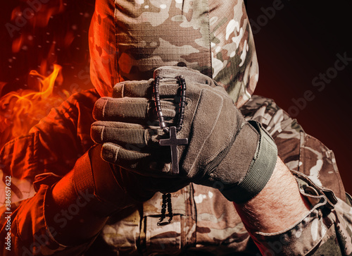 Photo of a military soldier in uniform and armor helmet sitting and holding cross religious necklace on back background with red fire burning glow. photo