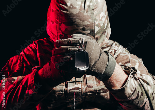 Canvas Print Photo of a military soldier in uniform and armor helmet sitting and holding metal army necklace on back background with red glow