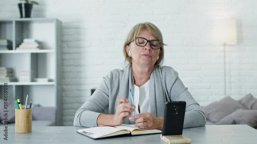 Woman Sitting At The Table And Talking To Someone On The Phone Via Video Link. She Works Remotely From Home Or Just Talks To Friends. She Makes Notes In Notebook. Woman Sitting At The Table And Talking To Someone On The Phone Via Video Link. She Works Remotely From Home Or Just Talks To Friends. She Makes Notes In Notebook.