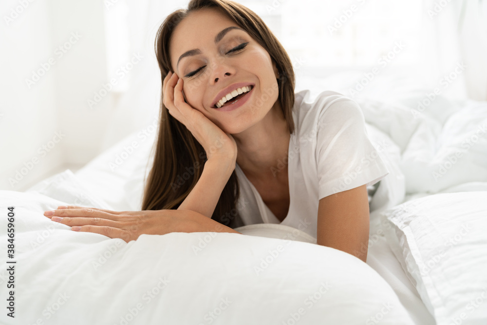 Fototapeta premium Happy weekend morning. Smiling millennial woman relaxing in white comfortable bed, waking up in a good mood, enjoy new day. Oral hygiene, wide toothy smile concept. 