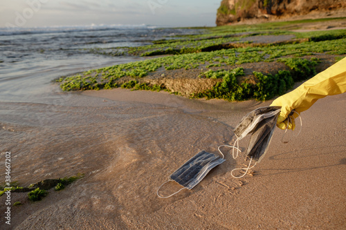 Problem of ocean polution after pandemia of covid. Female person in gloves picking up trash on the beach, medical face mask