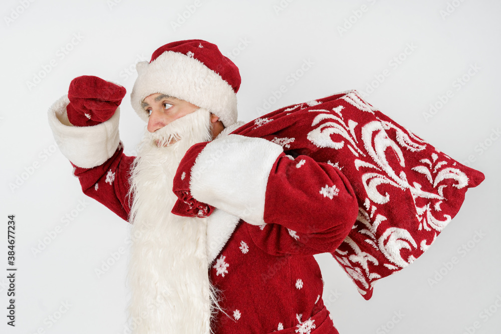 Santa Claus holds a bag of gifts over his shoulders and looks into the distance. He holds his hand to his forehead.