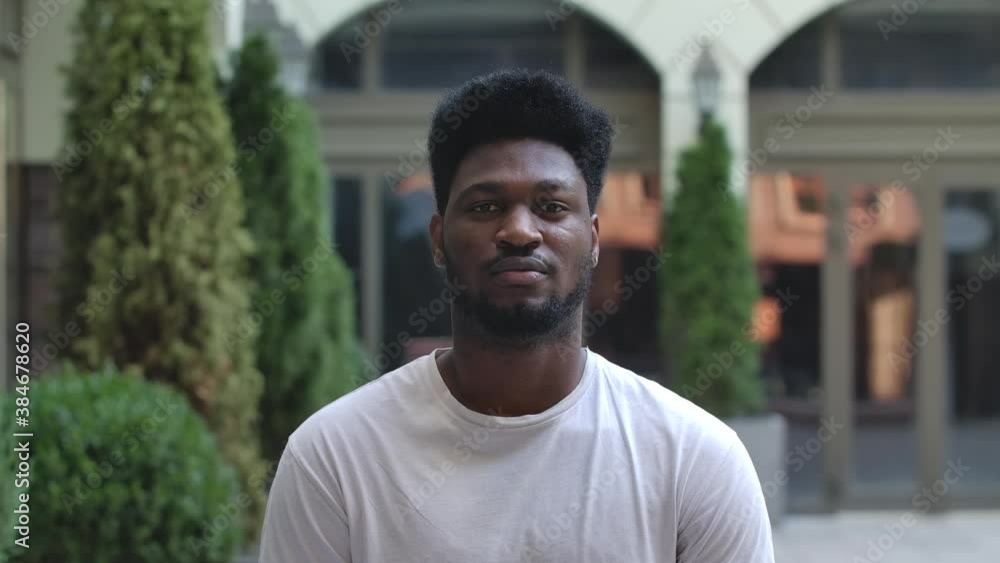 Portrait of young African American man looking at the camera. Stylish brunet in a white T shirt on the background of the street outdoors. Slow motion. Close up. Portrait of young African American man looking at the camera. Stylish brunet in a white T shirt on the background of the street outdoors. Slow motion. Close up.