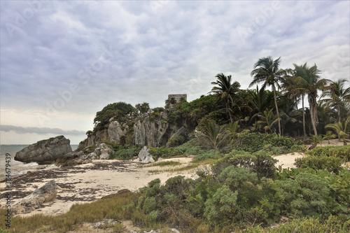 Beach on the Caribbean coast. A small sandy area surrounded by rocks and palm trees. At the top are the ruins of the ancient Mayan city of Tulum. Cloudy sky. Mexico.