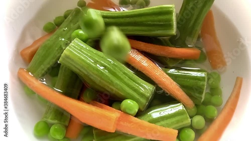 Dropping carrots drumstick pieces and fresh peas into a dish filled with water and vegetables