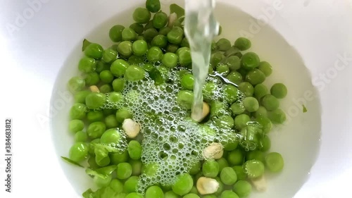 Pouring water and dropping drumstick pieces carrots and tomatoes into a dish of organic fresh green peas and spinach leaves