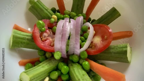 Dropping wet fresh organic green peas and corn kernels into a dish with exotic decoration of cut carrots onion rings tomato slices and drumstick pieces