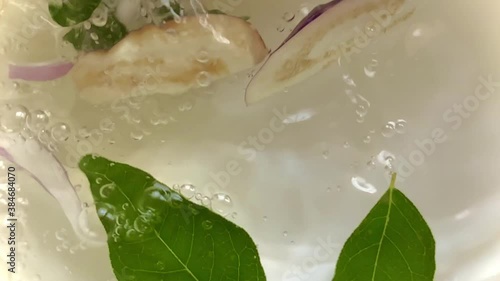 Pouring water and dropping chopped eggplant vegetable and onions slices into a bowl with leaves and green chilies creating beautiful splashes bubbles and ripples