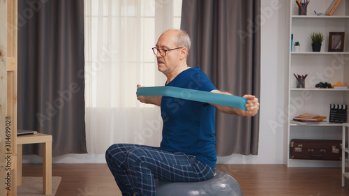 Strong senior man working out in living room using balance ball and resistance band. Old person pensioner healthy training healthcare sport at home, exercising fitness activity at elderly age © DC Studio