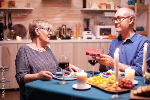 Senior man surprising wife with gift box during dinner. Happy cheerful elderly couple dining together at home  enjoying the meal  celebrating their anniversary  surprise holiday