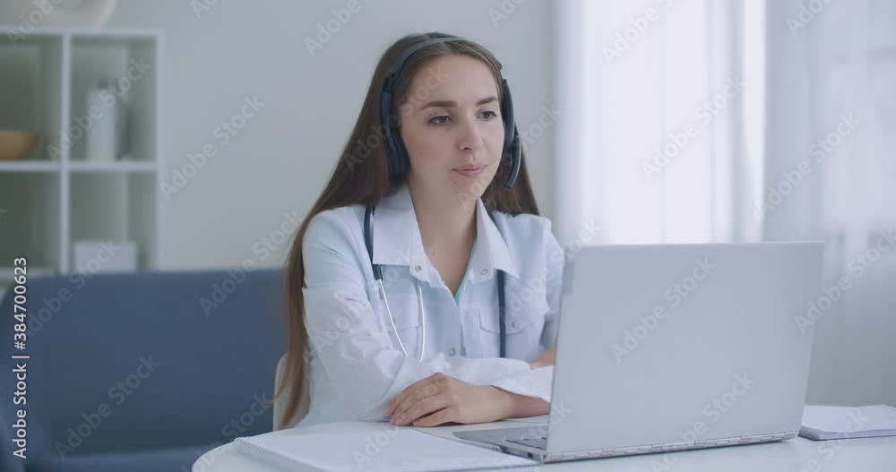 Female medical assistant wears white coat, headset video calling distant patient on laptop. Doctor talking to client using virtual chat computer app. Telemedicine, remote healthcare services concept. Female medical assistant wears white coat, headset video calling distant patient on laptop. Doctor talking to client using virtual chat computer app. Telemedicine, remote healthcare services concept.