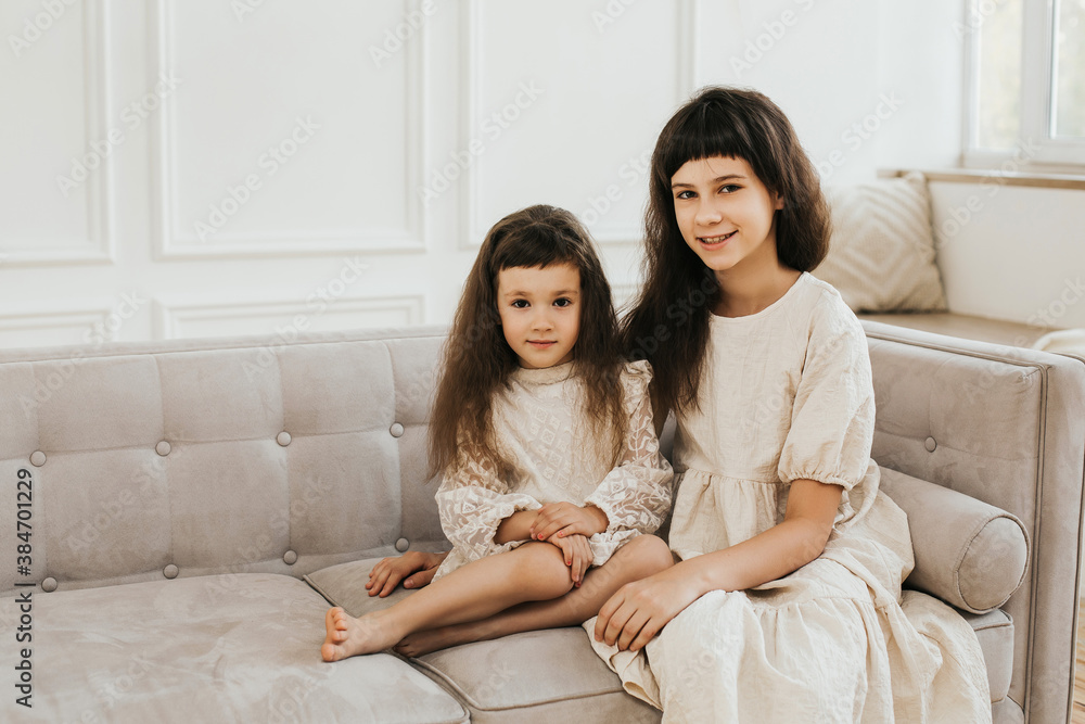 Two sisters are sitting on a sofa in a beautiful Scandinavian interior. Lovely little girls. Portrait of two dark-haired children.