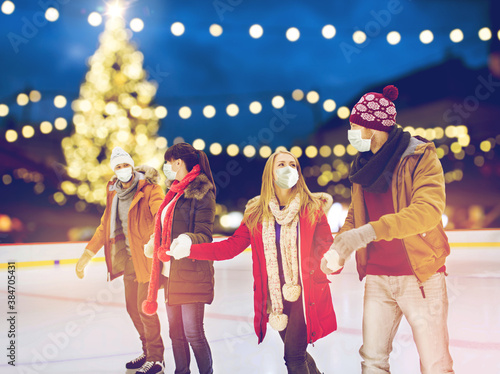 christmas, winter and leisure concept - friends wearing face protective medical masks for protection from virus disease holding hands at outdoor skating rink over holiday lights background